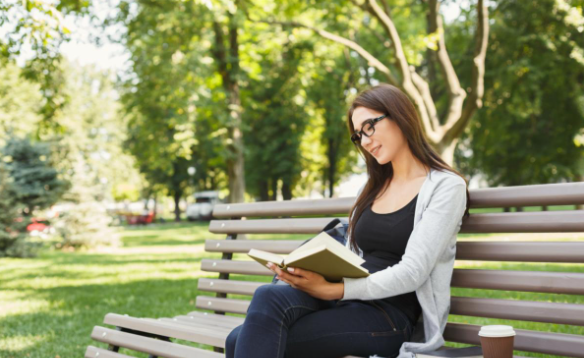 a young woman sitting on the bench in a park reading a book